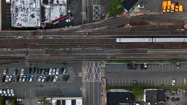 A top down, aerial view of a Long Island Railroad train passing by through an industrial area on Long Island on a cloudy day. The camera is tilted straight downward and remains stationary.