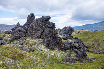 Rocky volcanic cliffs in Icelandic landscape surrounded by lush green moss and rolling mountains under cloudy skies in a picturesque natural setting. Basalt rocks