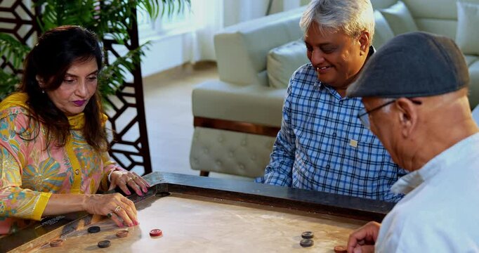 Indian seniors playing carrom board game together, sitting at dining table in cozy home interior, enjoying friendly indoor match while laughing, retired Indian men and women celebrating fun reunion ac