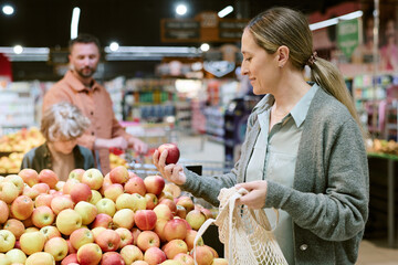 Woman selecting apples while holding net shopping bag. Background showing other shoppers and produce in well-lit grocery store