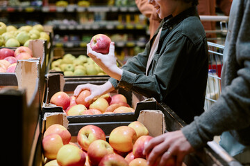 Individual choosing fresh apples in grocery store while holding one up for inspection. Shelves filled with a variety of apples surround person, showing abundant produce