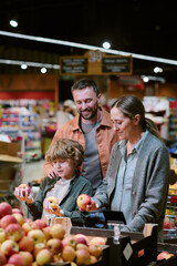Family standing in produce section selecting fresh apples together, enjoying grocery shopping with...