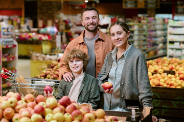 Family buying fresh apples at grocery store, standing by produce section and smiling for camera. Portrait of family enjoying quality time together in supermarket