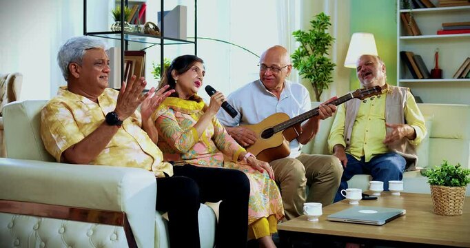 Seniors singing while playing guitar seated on comfortable sofa, Indian Asian elderly group of four friends reuniting joyfully indoors, enjoying musical bonding and happy moments at modern cozy home