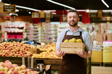 Grocer holding wooden basket filled with fresh yellow apples while standing in produce section of grocery store and smiling. Shelves stocked with various fruits in background