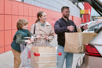 Family of three, including parents and child, placing grocery bags into car trunk while standing by shopping cart in outdoor parking lot © AnnaStills