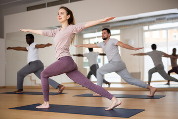 Obraz premium Motivated young woman maintaining active sporty lifestyle, training with group in yoga studio, standing in lunging asana Parivrtta Ashtachandrasana