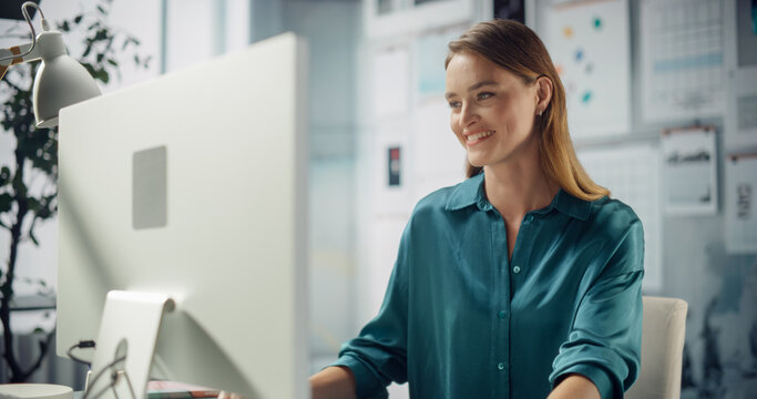 Woman in Teal Shirt Sits at Her Workspace, Using Computer. She Rests Her Chin on Her Hand, Smiling Thoughtfully. Background Features Screens and Creativity. Stylish Shot