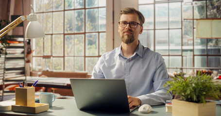 Bearded Professional Works on Laptop in Sunlit Office. Creative Man Looking at the Camera while Working from Home Office. Stylish Bright and Sunny Interior with Park in the Window. Medium Portrait