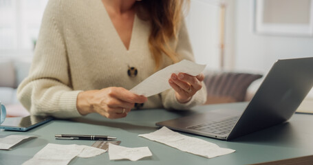 White Woman in Cream Cardigan Reviews Receipts at Her Laptop. Softly Lit Room With Bookshelves, Woman Does Taxes and BIlls, Organizing Financial Documents, Checks. Honest Taxpayer