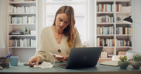 White Woman in Cream Cardigan Reviews Receipts at Her Laptop. Softly Lit Room With Bookshelves,...