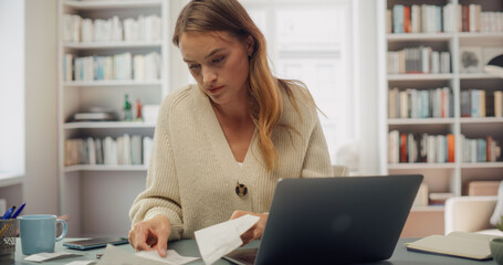 White Woman in Cream Cardigan Reviews Receipts at Her Laptop. Softly Lit Room With Bookshelves,...