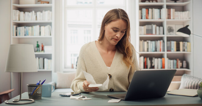 White Woman in Cream Cardigan Reviews Receipts at Her Laptop. Softly Lit Room With Bookshelves, Woman Does Taxes and BIlls, Organizing Financial Documents, Checks. Honest Taxpayer