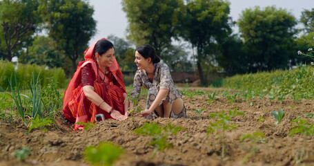 Indian active mother wear saree teen age cute girl sitting help and protect to grow tree in red soil young adult farmer female woman with lady talk gossip do nature care enjoy day desi life garden