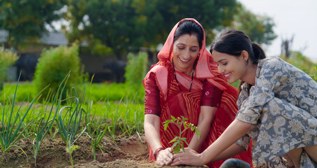 Indian active mother wear saree teen age cute girl sitting help and protect to grow tree in red soil young adult farmer female woman with lady do nature care enjoy day desi life garden