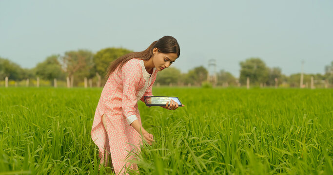 Indian young adult farmer woman lady wear casual dress desi life outdoor holding tablet device website looking screen asian smiling teen age girl use tab yield data outside work rural area day time - Powered by Adobe