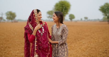 Indian asian villager farmer woman lady wear red saree smiling standing outdoor farmland having funny happy mom teen age young adult girl daughter talking gossip enjoy desi life day time outside	