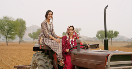 Indian village farmer mature woman lady wear red saree smiling sitting on tractor looking at camera outdoor asian mother with teen age girl daughter do pose staring cam outside land © Raushan_films