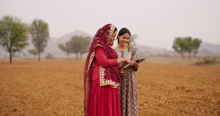 Fototapeta premium Indian asian villager farmer young adult woman lady wear red saree smiling outdoor hold tab device browse website data happy teen age girl teach mom talking enjoy day time outside desi life