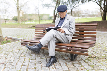Fototapeta premium Senior man reading a book and listening to music on a park bench