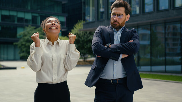 Happy successful smiling celebrating win African American woman businesswoman female in city joyful Caucasian man male businessman offended jealous envy different emotions two business colleagues