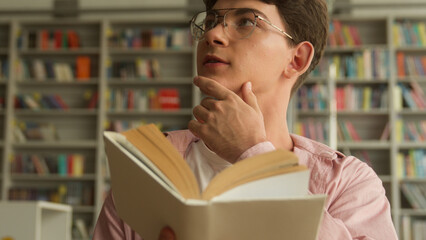 Caucasian teenager guy reading book amazed open mouth insight thinking preparing exams study in school college library teen boy read literature education studying learning homework think thoughtful