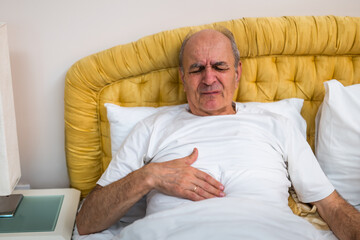 Distraught senior man suffering from stomachache while lying in bed at his home.