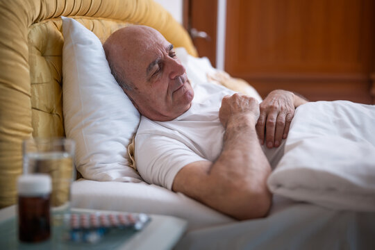 Senior man calmly sleeping in bed with pills and glass of water next to him on night table in his bedroom.