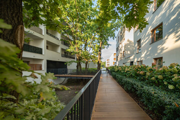 The well-landscaped inner territory of a modern residential complex. The recreational area features a wooden boardwalk, a bridge over a ditch, and lush greenery between buildings.