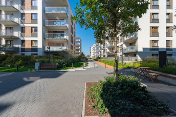 Wide pedestrian promenade in a new residential housing estate. This large public space includes various seating areas with benches and tables, surrounded by landscaped greenery and buildings.