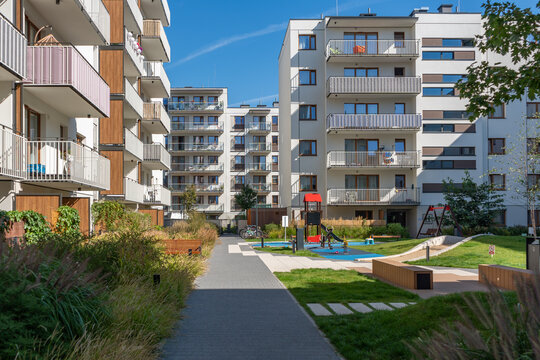 Multifunctional community space in a new residential development. The landscaped courtyard features a children's playground, wooden benches, a green lawn, and a pedestrian walkway.