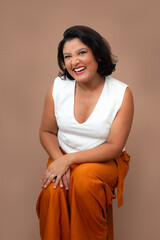 Cheerful businesswoman in white top and orange pants sitting on a stool and laughing at camera. Perfect for happiness at work, corporate well-being, friendly approach. Studio portrait with copy space.