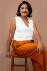 Elegant businesswoman in white top and orange pants sitting on a stool, looking at camera. Perfect for professional development, career, personal growth. Copy space on mocha mousse color background.