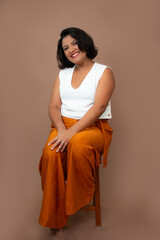 Charming businesswoman in white top and orange pants sitting on a stool and smiling at camera. Ideal for happiness at work, corporate well-being, friendly approach. Vertical portrait with copy space.