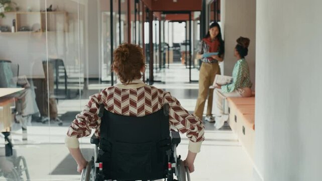 Rear tracking shot of male redhead office worker with disability riding in wheelchair along light corridor, coworkers talking, inclusivity, mobility and equal opportunities in creative workspace
