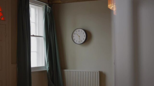 A room with a clock on the wall and a window with green curtains. The room is empty and the clock is set to the time of 10:00