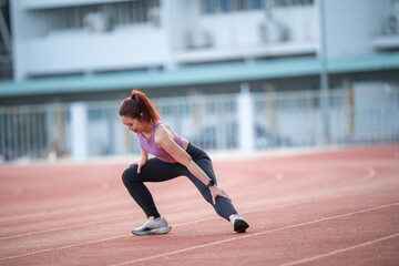 A woman is stretching on a track. She is wearing a purple shirt and black pants. She is bending over and reaching her leg