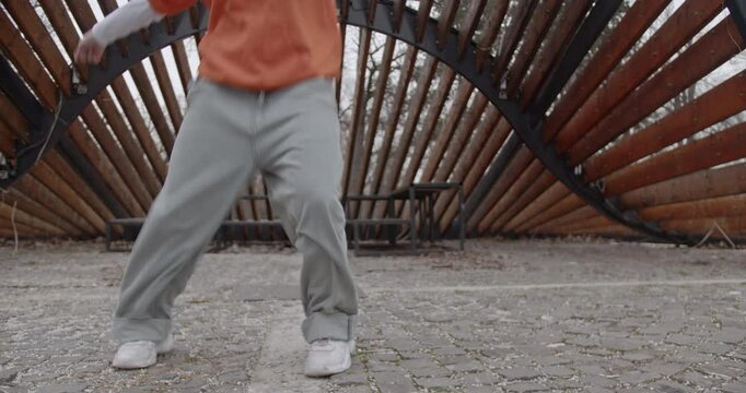 Young woman dancing contemporary or modern dance choreography in an urban park setting. She's wearing baggy gray pants and an orange hooded sweater.