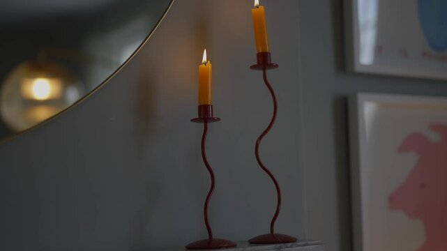Two candles are lit and are standing on a red stand. The candles are yellow and are glowing brightly. The stand is made of metal and has a curved design. The room is dimly lit, creating a cozy