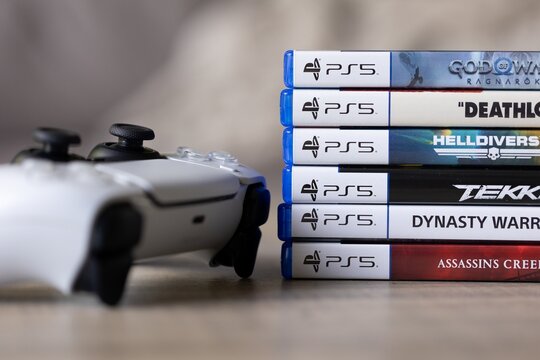 Brecht, Belgium - 18 april 2025: A stack of sony playstation 5 videogame cases or boxes lying on a wooden table next to a wireless PS5 controller. Ready to play a console game.