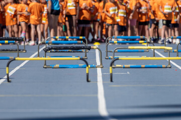 Blue athletics track with yellow and black hurdles aligned and young athletes waiting in the background