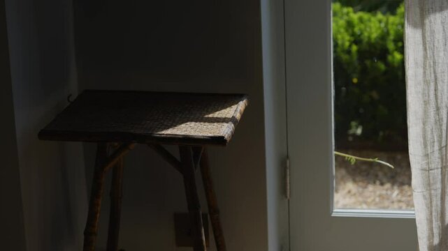 A wooden table with a wicker top sits in front of a window. The window is open, letting in sunlight and a view of the outside. The table is empty