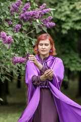 A beautiful young woman with red hair, against a background of blooming lilacs, in a cloak and hood. Medieval fantasy costume. Fairy tale heroine, princess, duchess.