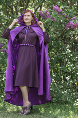 A beautiful young woman with red hair, against a background of blooming lilacs, in a cloak and hood. Medieval fantasy costume. Fairy tale heroine, princess, duchess.