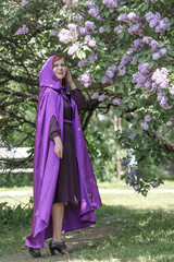 A beautiful young woman with red hair, against a background of blooming lilacs, in a cloak and hood. Medieval fantasy costume. Fairy tale heroine, princess, duchess.