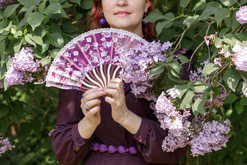 A beautiful young woman with red hair, against a background of blooming lilacs, in a cloak and hood. Medieval fantasy costume. Fairy tale heroine, princess, duchess.