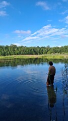 Fototapeta premium Tranquil lake scene with clear blue water, green reeds, and a lush forest under a bright sky. Perfect nature landscape for relaxation, travel, or outdoor themes.