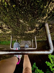 Person climbing a rusty ladder on a moss covered wall surrounded by lush greenery during daylight