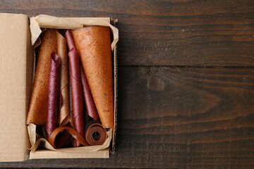 Delicious fruit leather rolls in cardboard box on wooden table, top view. Space for text