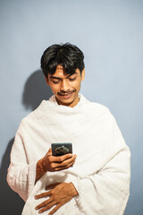 A young Muslim man dressed in traditional white ihram clothes is smiling while looking at his...
